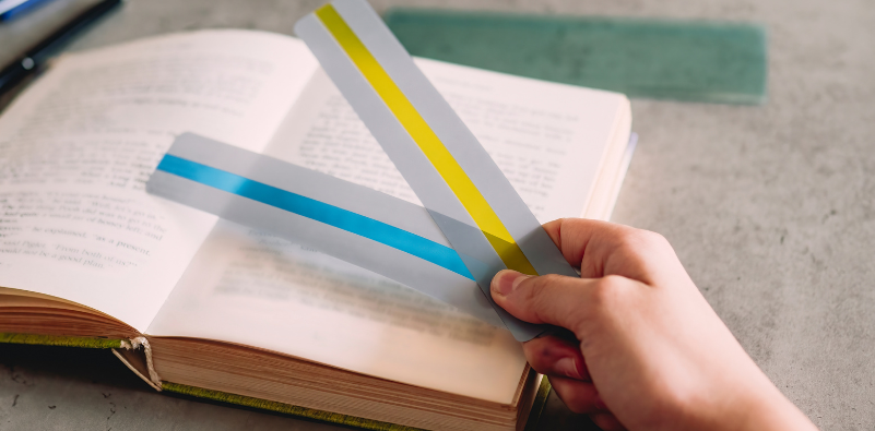 A child holding blue and yellow coloured overlays in their hand over a book.