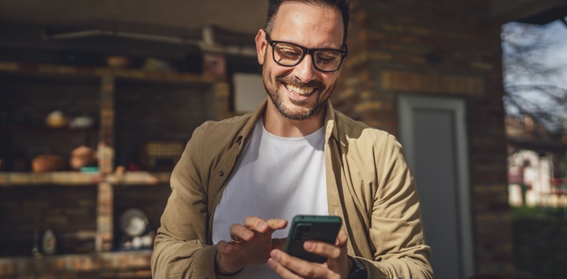 A man with a beard and glasses standing outside using a smart phone.