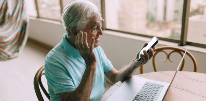 An old man on the phone and laptop touching his ear.