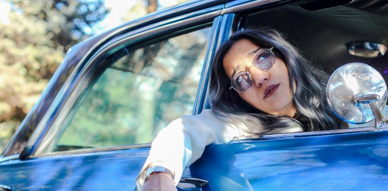 Woman in a blue car wearing blue tinted sunglasses looking outside the window.