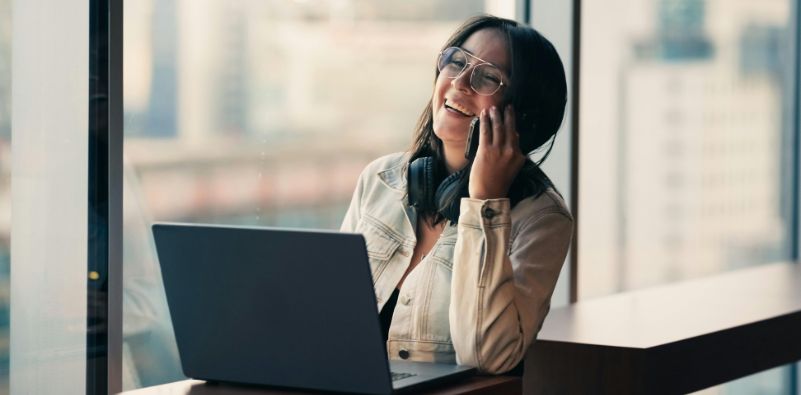 Woman in an office setting with a laptop on th desk and her laughing on the phone