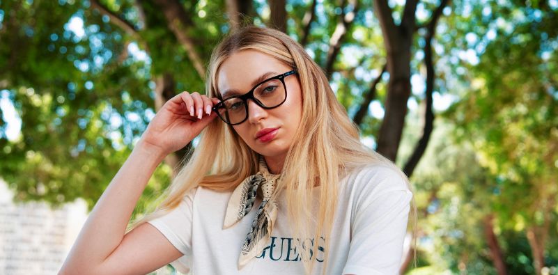 A woman sat under the trees wearing glasses and a white t-shirt